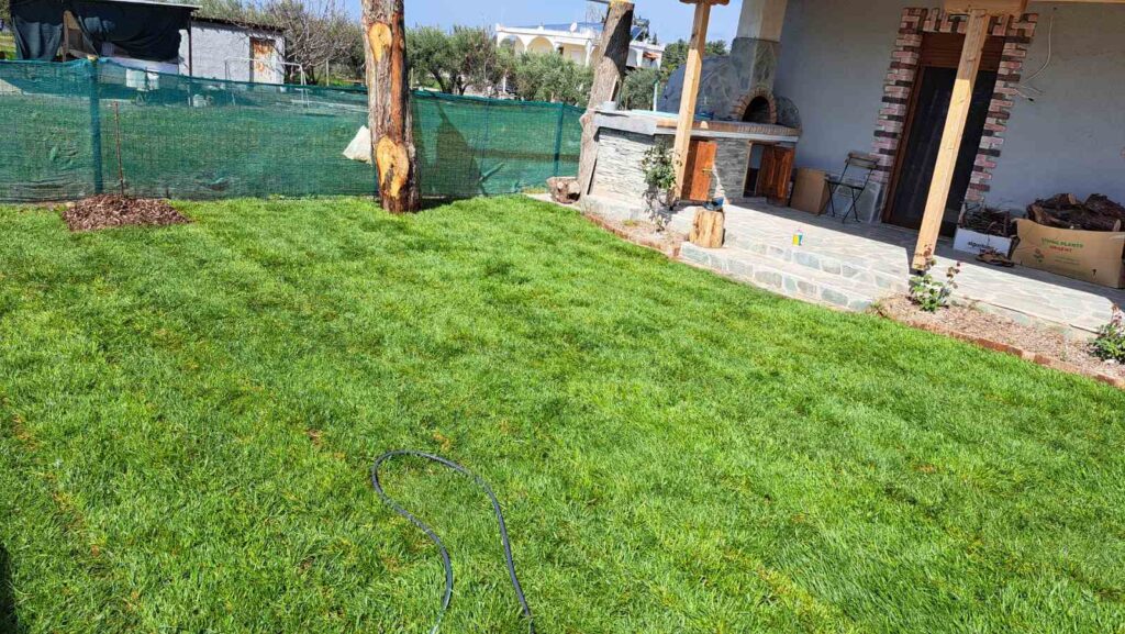 Backyard with lush green grass, a stone patio, and a brick outdoor kitchen under a covered area; a green construction fence in the background.