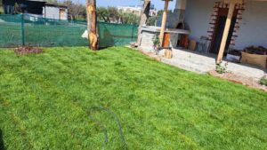 Backyard with lush green grass, a stone patio, and a brick outdoor kitchen under a covered area; a green construction fence in the background.