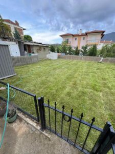 Backyard with a well-kept green lawn, a black metal fence, and a hose across the grass; pink houses and trees in the background.