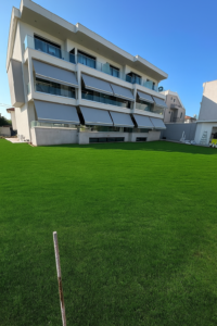 Modern white apartment building with staggered balconies and glass railings beside a well-kept green lawn, under a blue sky.