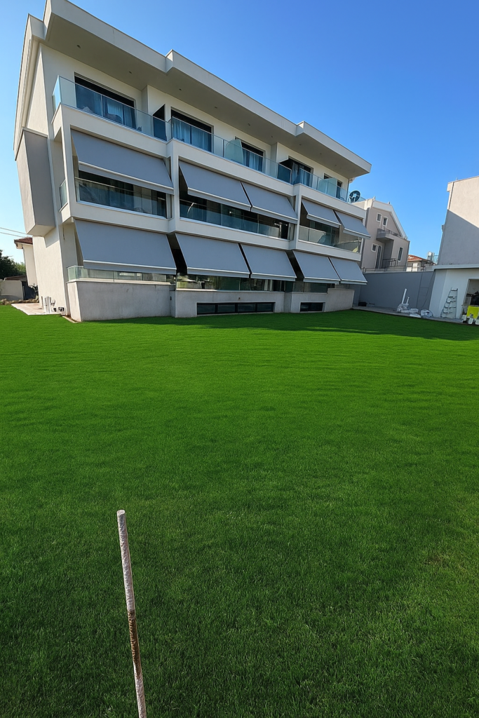Modern white apartment building with staggered balconies and glass railings beside a well-kept green lawn, under a blue sky.