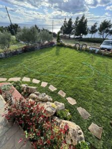 A well-kept lawn with stepping-stone path near a rocky flower bed, overlooking the sea and trees in the background; a garden hose lies across the grass.