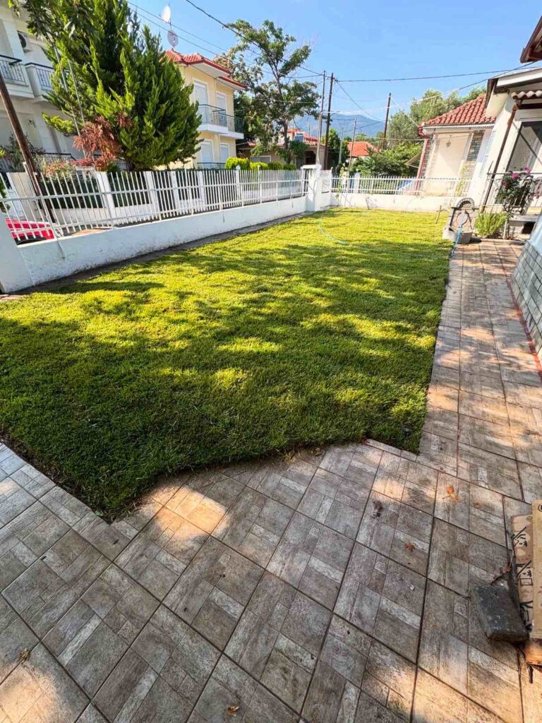 Sunlit suburban yard with a lush green lawn bordered by a white fence and a gray tiled patio along the right side.