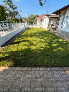 Sunlit front yard of a house with a green lawn, white fence along the left, and a tiled path on the right side.