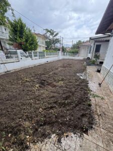 A small backyard with bare soil ready for planting, bordered by a white fence and paved walkway on the right, with neighboring houses and trees beyond.